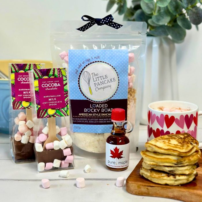 A rocky road pancake mix pack, two hot chocolate spoons, a mini bottle of maple syrup and a stack of pancakes with a mug of hot chocolate and plant in the background.