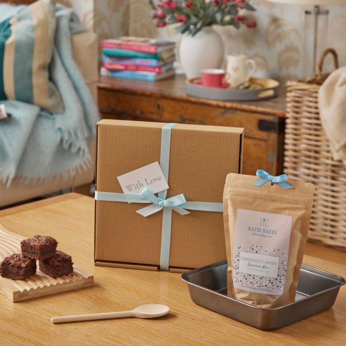 gift box tied with blue ribbon ona a table with a brownie tin, baked brownies, a brownie baking mix and a wooden spoon.