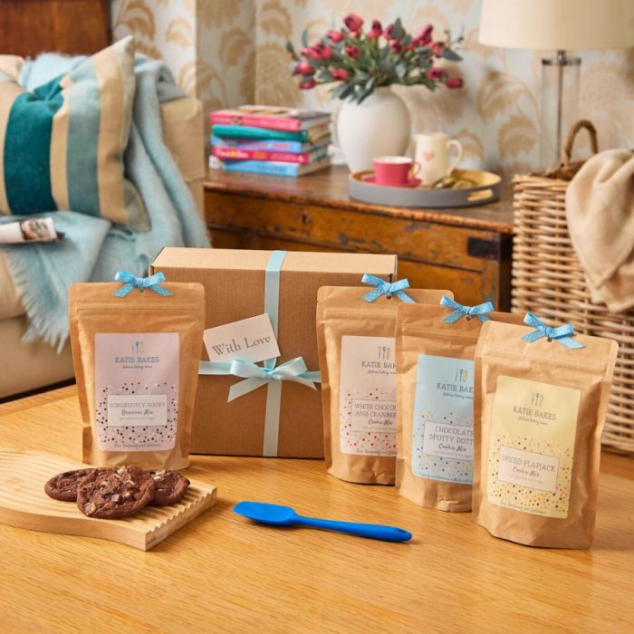 Hamper gift box tied with blue ribbon on a table with 4 baking mix pouches, a silicone spoon and a board with baked cookies.