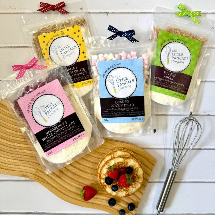 The contents of a Pancake Lovers gift hamper with Four gourmet pancake mixes and a whisk with a stack of baked pancakes and fresh berries