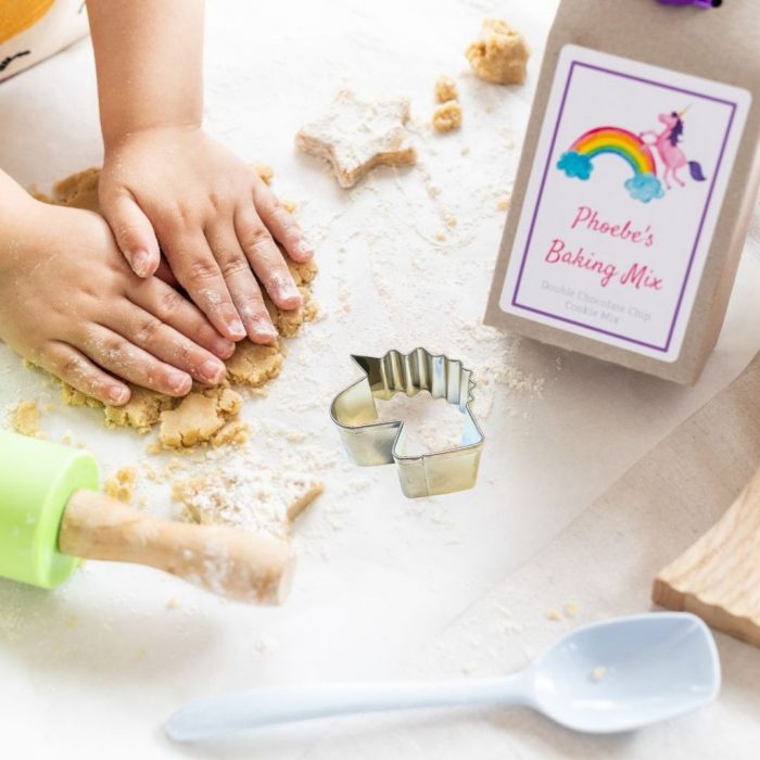 Cookie mix inside a gift box with an image of Rainbows and Unicorn and text of your choice on the box with a purple ribbon at the top. A stainless steel cutter in the shape of a Unicorn. The mix is being made up in the background showing a rolling pin and spoon.