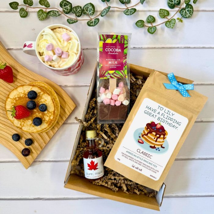 A breakfast-in-bed gift set arranged on a white wooden surface. The set includes a pink mug filled with hot chocolate, whipped cream, and mini marshmallows; a wooden board with pancakes topped with strawberries and blueberries; a small bottle of pure Canadian maple syrup; a packet of pancake mix with a personalized label that reads "To Lily, Have a flipping great birthday"; and a marshmallow milk hot chocolate spoon by Cocoba. The items are placed in a kraft paper box with decorative shredded paper and a blue ribbon, with green vine decorations nearby.