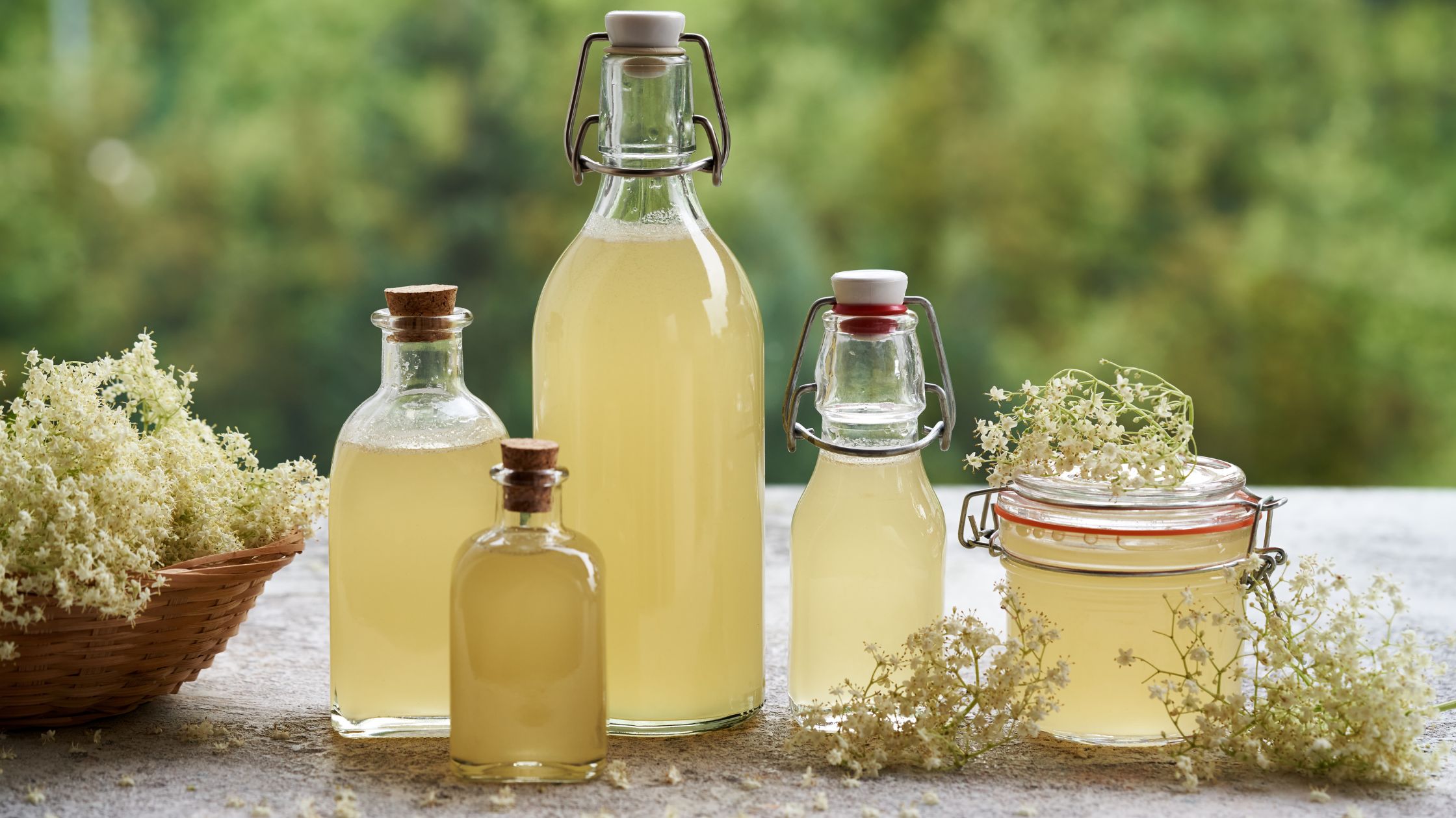 A selection of sterilised glass bottles and jars filled with homemade elderflower cordial, surrounded by fresh elderflowers on a rustic table with a blurred green background—perfect for illustrating an elderflower cordial recipe with emphasis on sterilisation and natural ingredients.