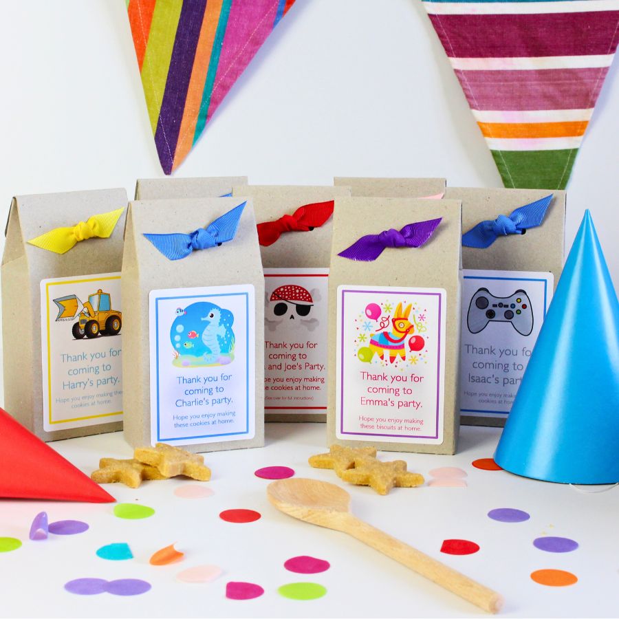 Eco-friendly, award-winning personalised kids' party bags by Katie Bakes, featuring bidegradable packaging with colourful ribbons and custom thank-you labels. Each party bag includes a fun baking kit, ideal for children's birthday party giveaways. Designs feature themes like dinosaurs, diggers and trucks, pirates, piñatas, and video games. Surrounded by festive party decorations including confetti, wooden spoon, party hats, and bunting.