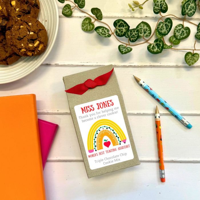 A gift package labeled "MISS JONES" with a red ribbon and a colorful tag that reads "Thank you for helping me become a clever cookie! WORLD'S BEST TEACHING ASSISTANT, Triple Chocolate Chip Cookie Mix." The package is placed on a white wooden surface alongside two colorful pencils, two notebooks (one orange, one pink), a green trailing plant, and a plate of triple chocolate chip cookies.