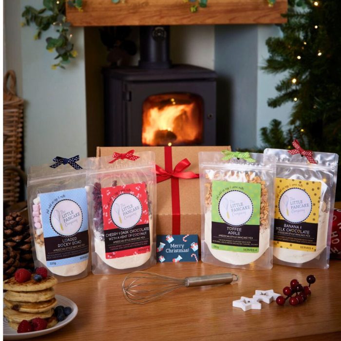 "A festive Christmas Pancake Lovers’ Gift Hamper from The Little Pancake Company displayed on a wooden table in front of a glowing wood-burning stove and decorated Christmas tree. The hamper includes four pancake mix pouches in flavors Loaded Rocky Road, Cherry & Dark Chocolate with a Hint of Coconut, Toffee Apple, and Banana & Milk Chocolate, each topped with a small ribbon matching its label. Behind them sits a brown gift box tied with a red ribbon and a 'Merry Christmas!' card. A whisk, pinecones, stacked pancakes topped with fresh berries, and festive decorations complete the cozy holiday scene.