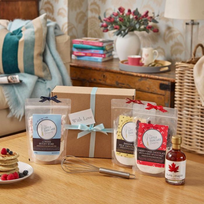 Hamper box tied with ribbon sitting on a table with three gourmet pancake mixes, bottle of maple syrup and whisk.