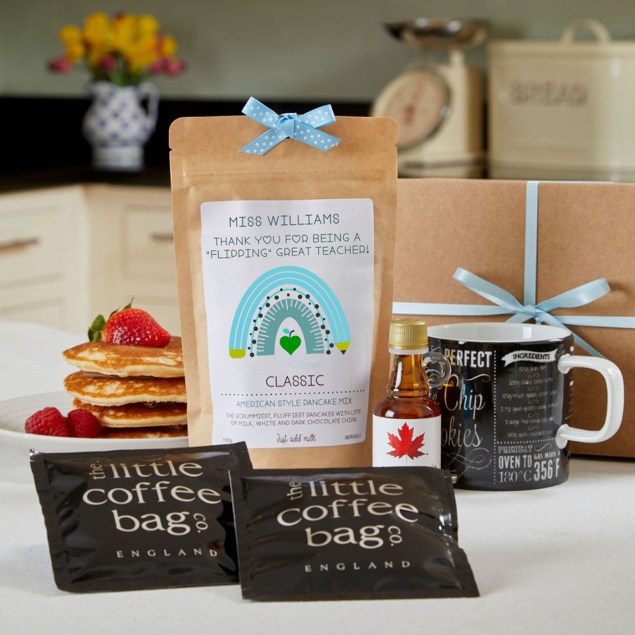 Gift set for a teacher displayed on a kitchen counter, featuring a personalized pancake mix pouch labeled "Miss Williams," two coffee bags, a bottle of maple syrup, a black cookie-themed mug, and a wrapped gift box with blue ribbon. A stack of pancakes with fresh strawberries and raspberries sits beside the gift, with a softly blurred kitchen background.